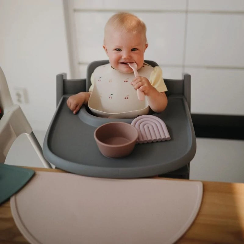 Bébé en chaise haute tenant une cuillère en silicone rose avec bavoir et bol assortis